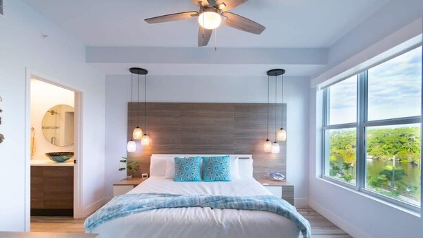 A bedroom with a bed, a ceiling fan, pendant lights, a window with a view, and a wooden headboard. - Waterfront Stateroom (Merritt Island)