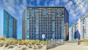 A high-rise building with a grid-like facade, a sandy foreground, and a clear sky.