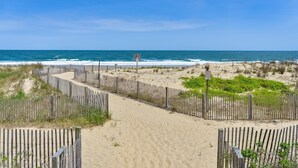 A sandy beach path with a wooden fence leading to the ocean, a sign indicating a dune crossing, and a clear view of the sea.