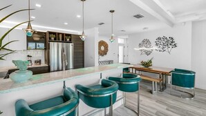 Moderni keittiö, jossa on keskeinen saareke, baarituolit, riippuvalaisimet ja koristeellinen seinätaide.