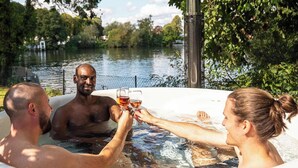 Un jacuzzi con tres personas brindando con bebidas, rodeado de árboles y una masa de agua.