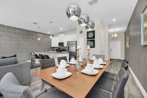 A modern dining area with a wooden table set for a meal, grey upholstered chairs, and a kitchen in the background.