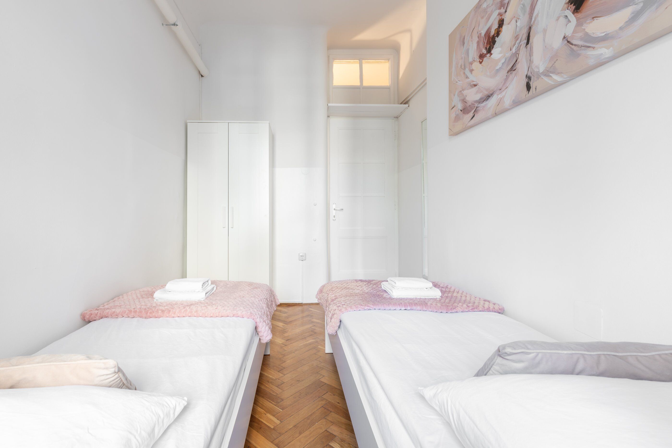 Une chambre d’hôtel avec deux lits simples, un parquet en bois disposé en point de Hongrie, des murs blancs et un tableau accroché au mur.