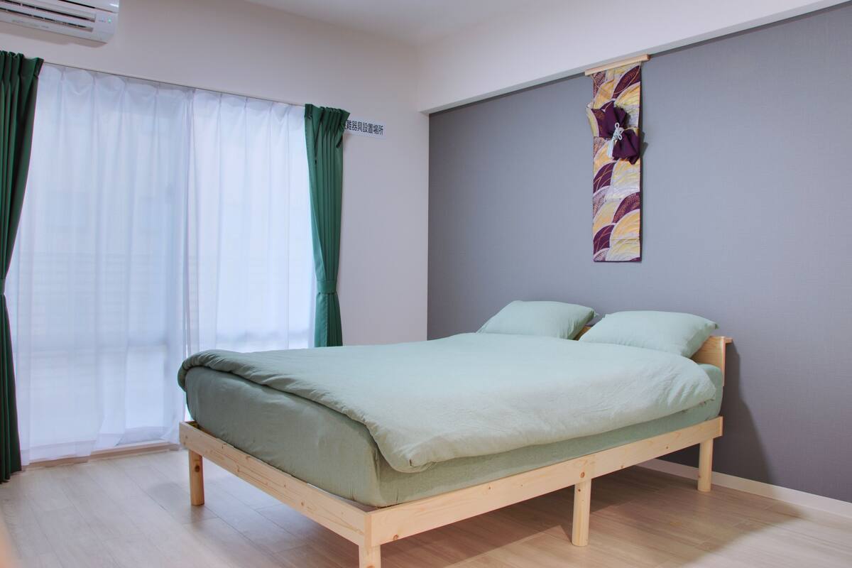 Un lit simple dans une chambre dotée d’un cadre en bois, d’un plancher de couleur claire et d’une fenêtre ornée de rideaux blancs transparents et d’un voilage vert.