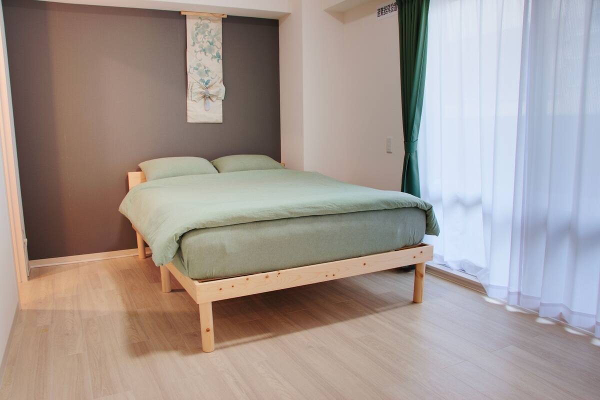 Un lit simple avec un cadre en bois et un matelas vert, dans une pièce dont le sol est recouvert de parquet clair et un des murs est peint dans une couleur foncée.