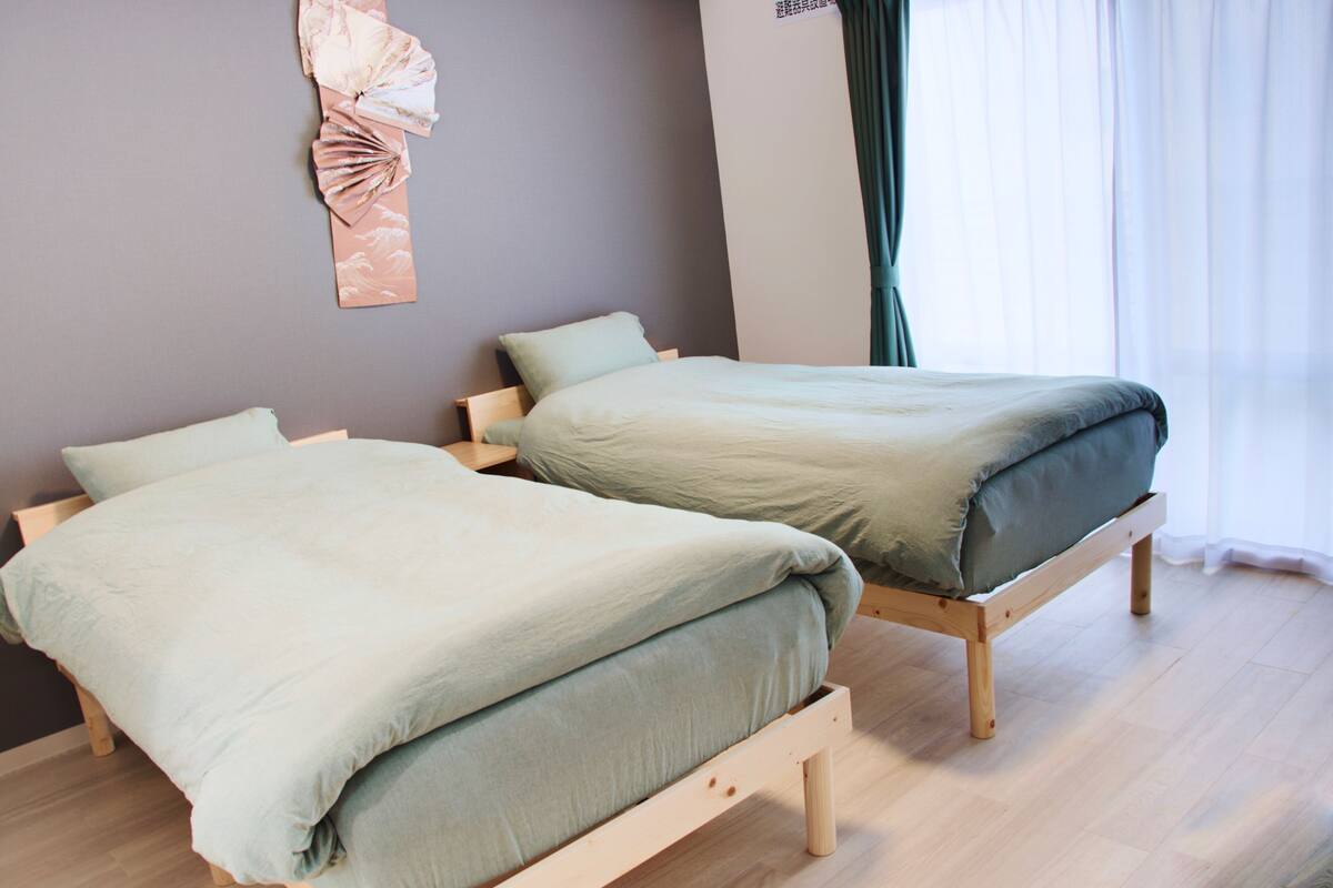 Deux lits simples avec des cadres en bois, dans une pièce aux murs de couleur claire et au plancher en bois.