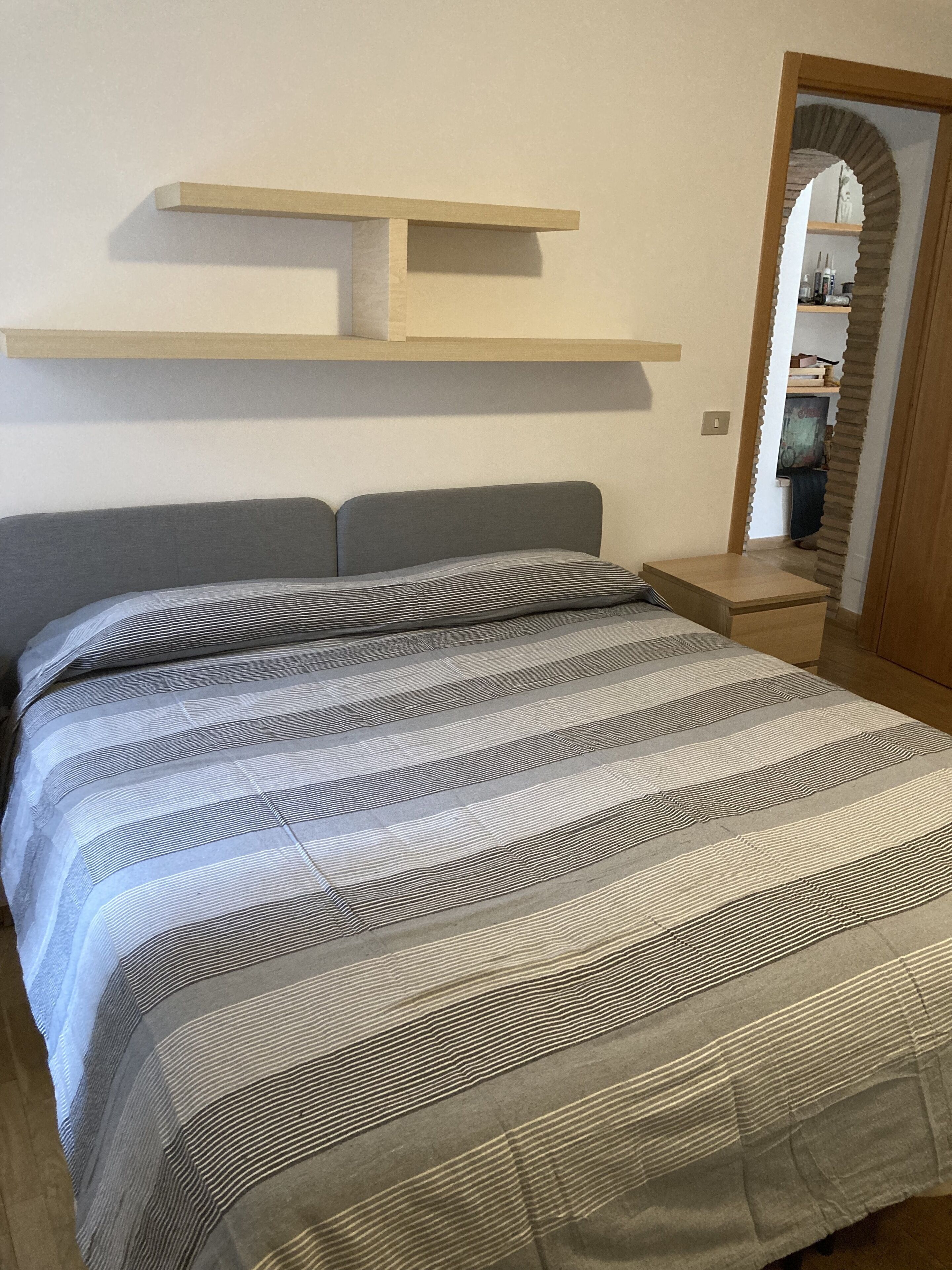 Ein Schlafzimmer mit einem Bett, einem Nachttisch, einem Regal über dem Bett und einem Spiegel mit Bogenrahmen.