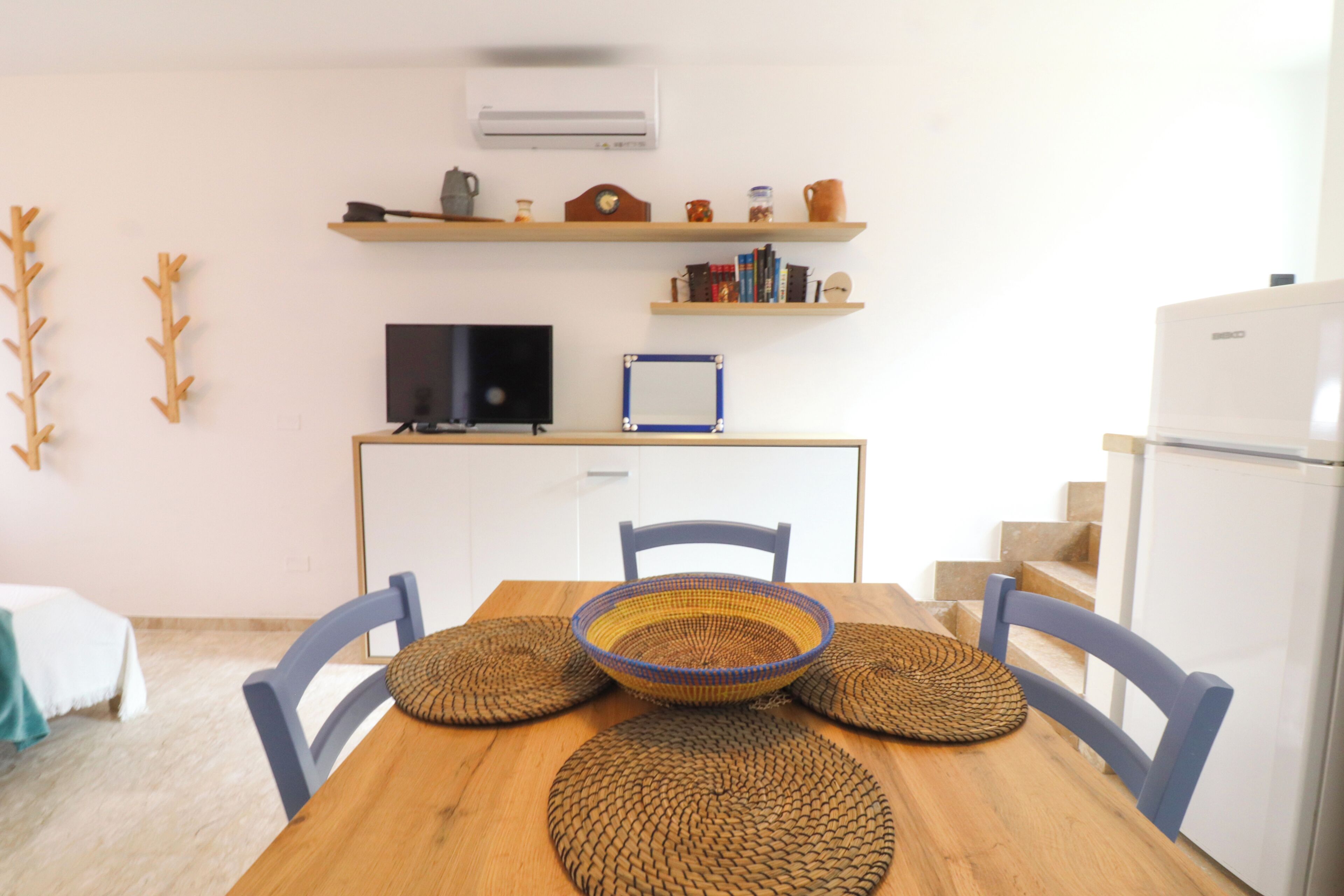 Un comedor con una mesa de madera, cuatro individuales tejidos, dos sillas, un televisor sobre un soporte y estantes sobre la encimera con varios objetos.