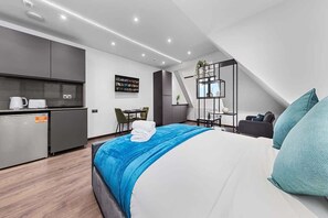 A modern hotel room with a large bed, a kitchenette, a dining area, and a small balcony.