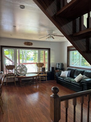 A room with a wooden floor, a ceiling fan, a wooden staircase, a green sofa, and a dining table with chairs.