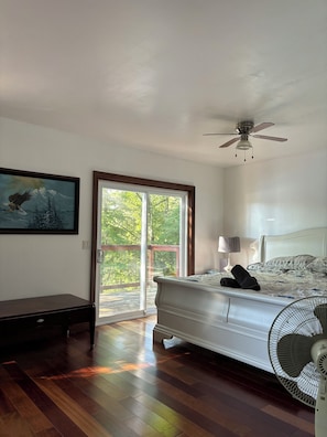 Une chambre à coucher comprenant un lit, un ventilateur de plafond, une table de chevet, une lampe, un tableau et une porte-fenêtre coulissante donnant sur une terrasse.
