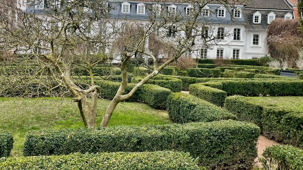 Un jardin bien entretenu, avec des haies taillées avec soin, et un grand bâtiment blanc doté de nombreuses fenêtres, visible au fond.