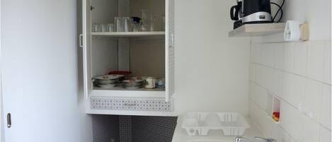 Una cocina con un armario blanco que contiene cristalería y vajilla, una cafetera en una repisa y un lavabo con grifo.
