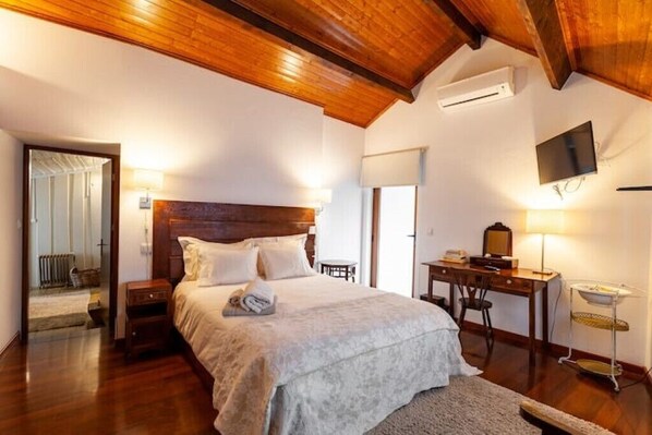 Un dormitorio con techo de madera, una cama con ropa de cama blanca, un escritorio con una silla, un televisor y un equipo de aire acondicionado.