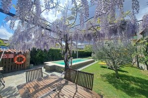 Una terraza de madera con piscina, rodeada de exuberante follaje y árboles de flores moradas.