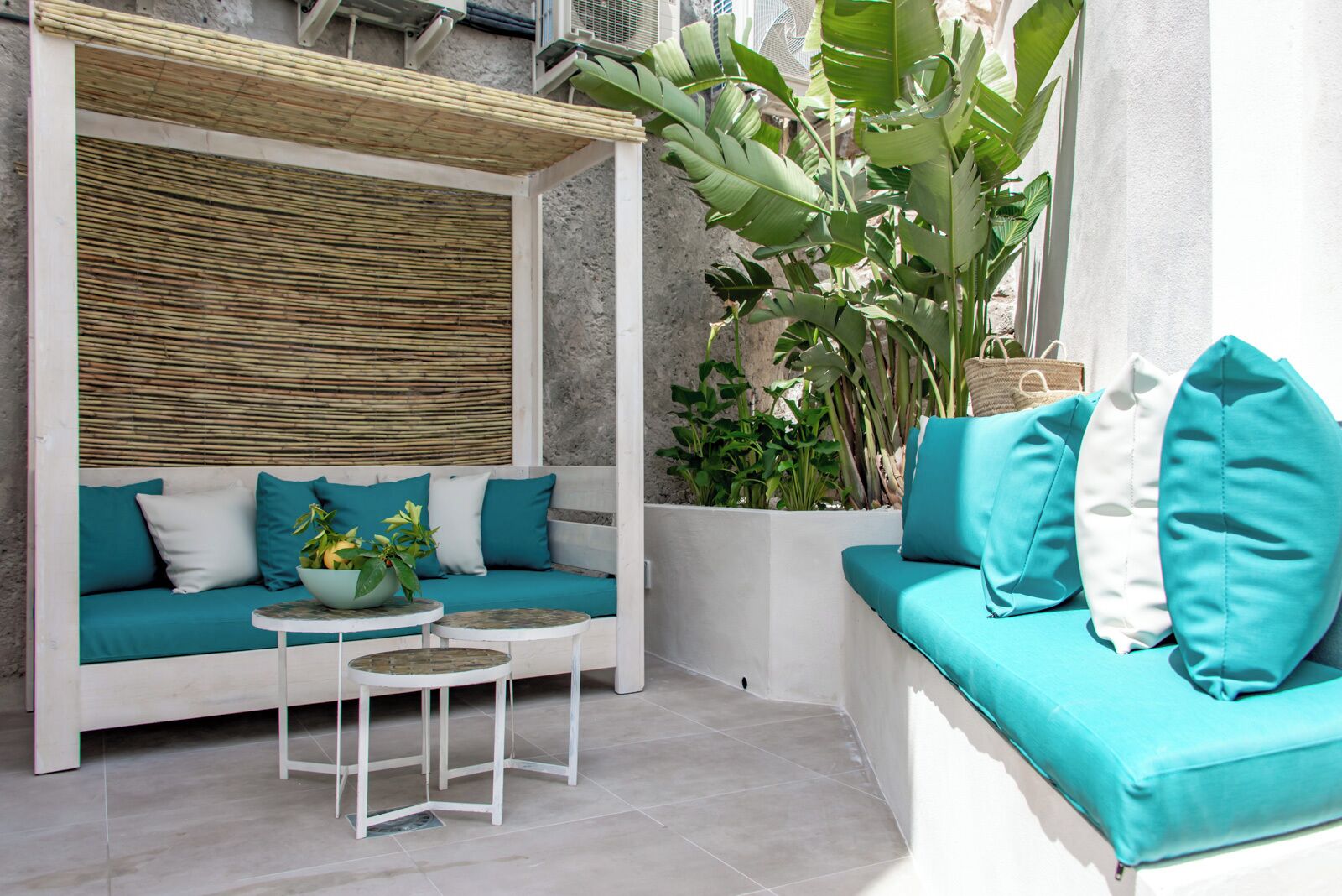 A modern outdoor seating area with a sofa and two small tables, surrounded by large green plants and a bamboo wall.