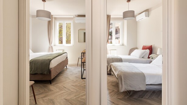 Reflejo de un cuarto de hotel con dos camas, piso de madera en espiga, un escritorio con silla y una ventana con cortinas.