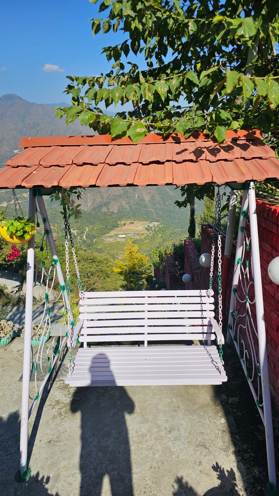 Un columpio con estructura metálica y techo rojo, rodeado de follaje y un paisaje montañoso al fondo.
