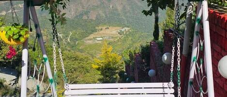 Un columpio con estructura metálica y techo rojo, rodeado de follaje y un paisaje montañoso al fondo.