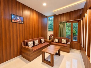 A modern living room with wooden paneling, a sectional sofa with patterned cushions, a coffee table, and a window with a view of greenery. - Hotel Luxmi Vilas Inn (Dhanaulti)