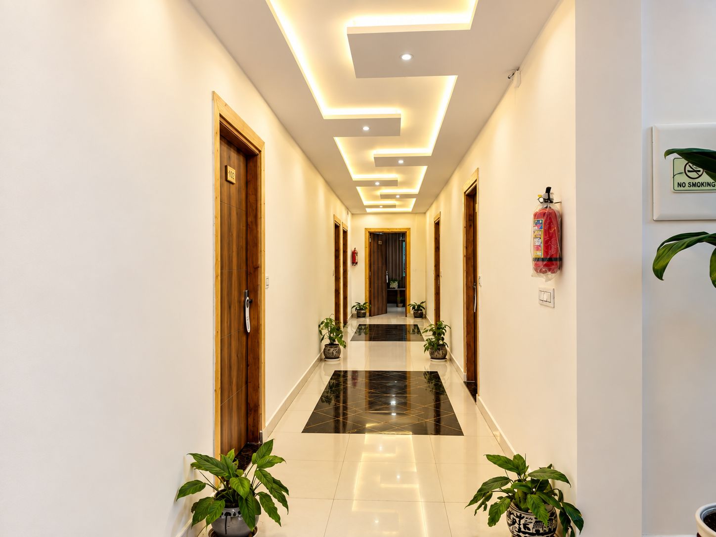 Un pasillo de hotel con diseño moderno, que presenta un suelo brillante, puertas de madera y plantas en macetas.