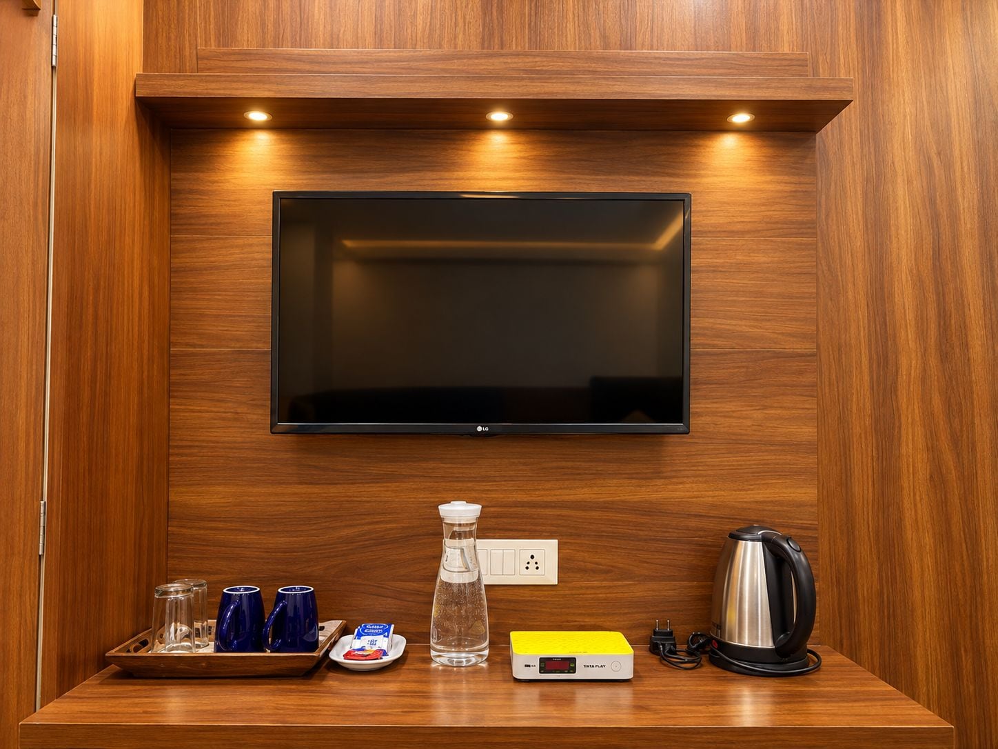 A hotel room with a flat-screen TV mounted on a wooden wall, a tray with glasses and a bottle, a kettle, and a digital clock.