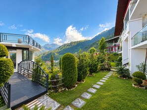 A modern building with a balcony, a stone pathway, and a well-maintained garden with various plants and a mountain view in the background. - Hotel Luxmi Vilas Inn (Dhanaulti)