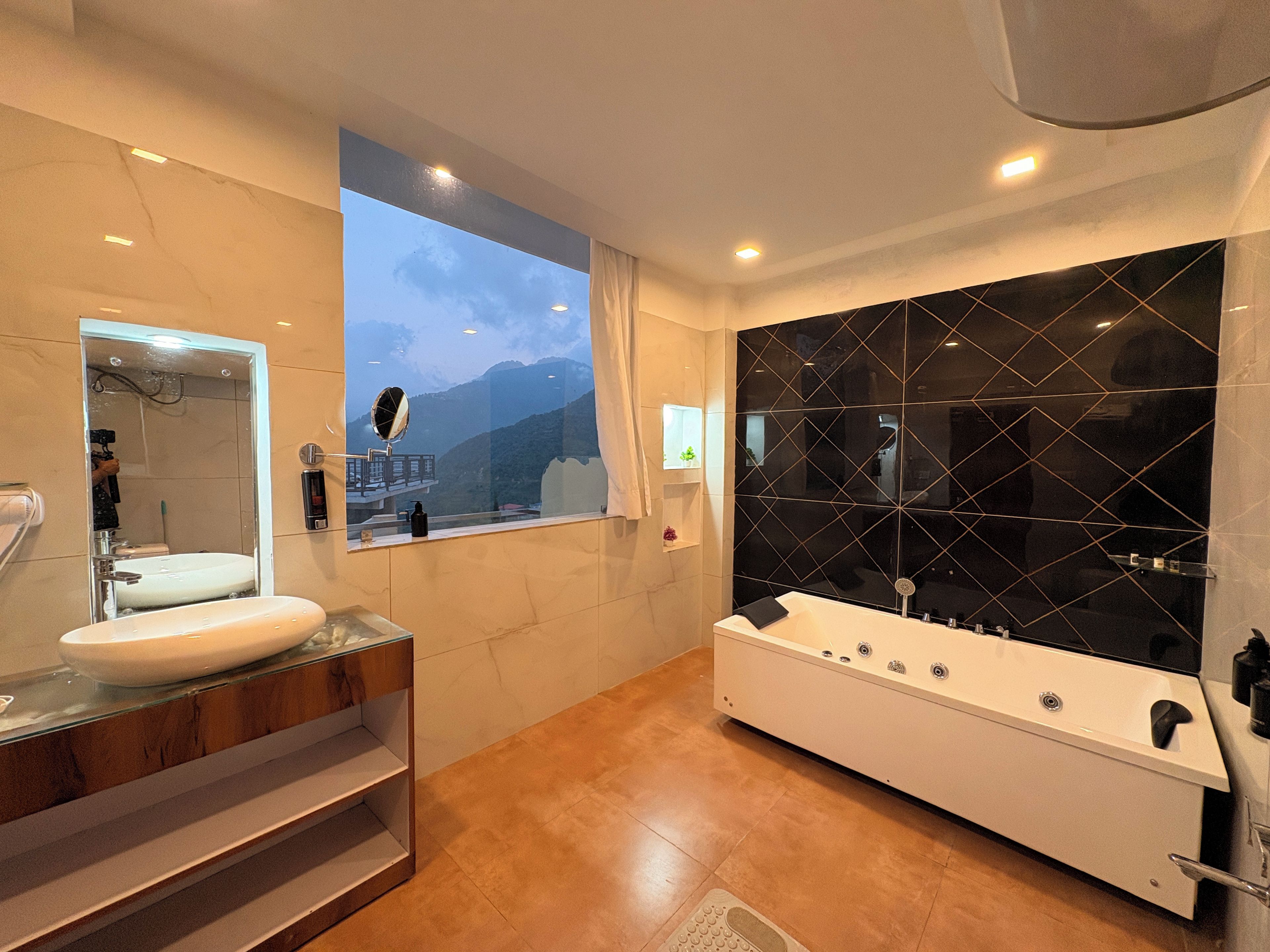 A modern bathroom with a large mirror, a bathtub, and a window with a view of mountains.
