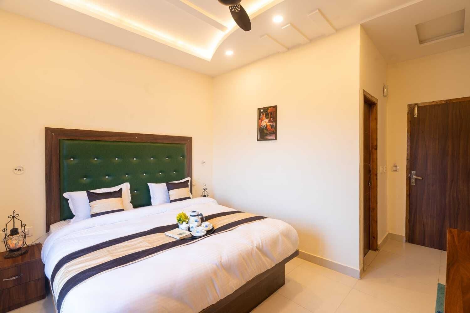 Een net opgemaakt bed met een groen hoofdeinde, een nachtkastje met een lamp, en een ingelijst foto aan de muur.