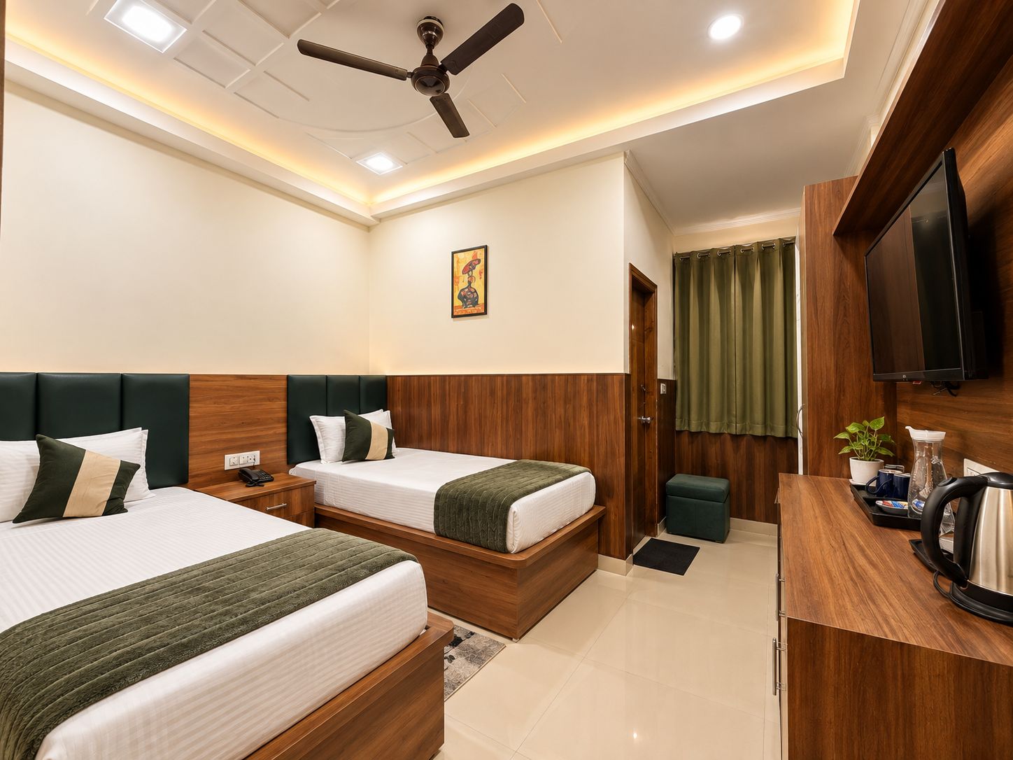 A hotel room with two beds, a ceiling fan, a flat-screen TV, a small kitchenette, and a painting on the wall.