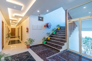 A modern hotel lobby with a marble staircase, potted plants, and a fire extinguisher on the wall.