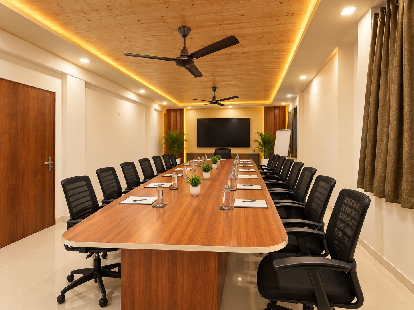 Un salón de conferencias con una larga mesa, sillas negras, un ventilador de techo y una pantalla grande.