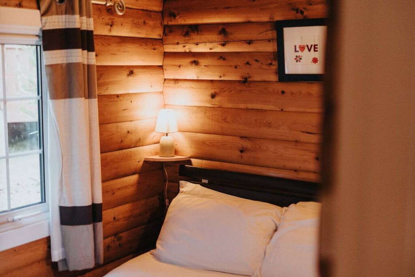 A cozy wooden room with a bed, a nightstand with a lamp, and a framed 'LOVE' sign on the wall.