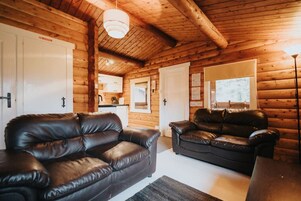 A log cabin interior with two leather sofas, a dining table, and a kitchen area.