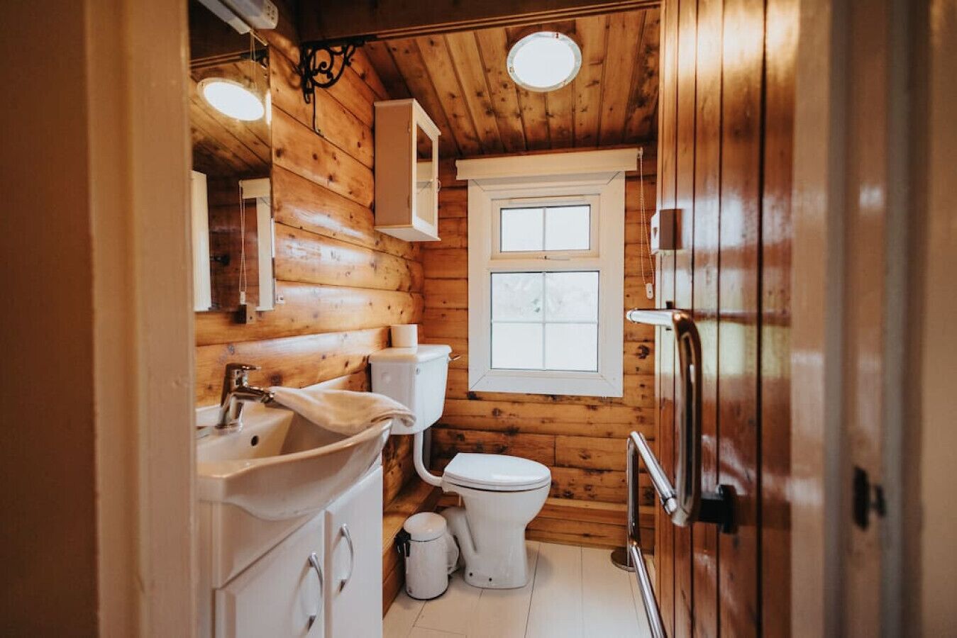 A compact wooden cabin bathroom with a sink, toilet, and window.