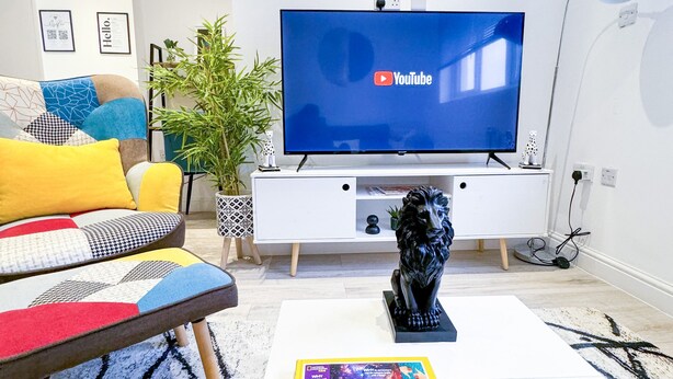 A modern living room with a flat-screen TV, a sofa with colorful cushions, a coffee table with a magazine, and a decorative lion statue. - Perfect 2 bed Apartment 4 Contractors Sleep Upto 5 (London)