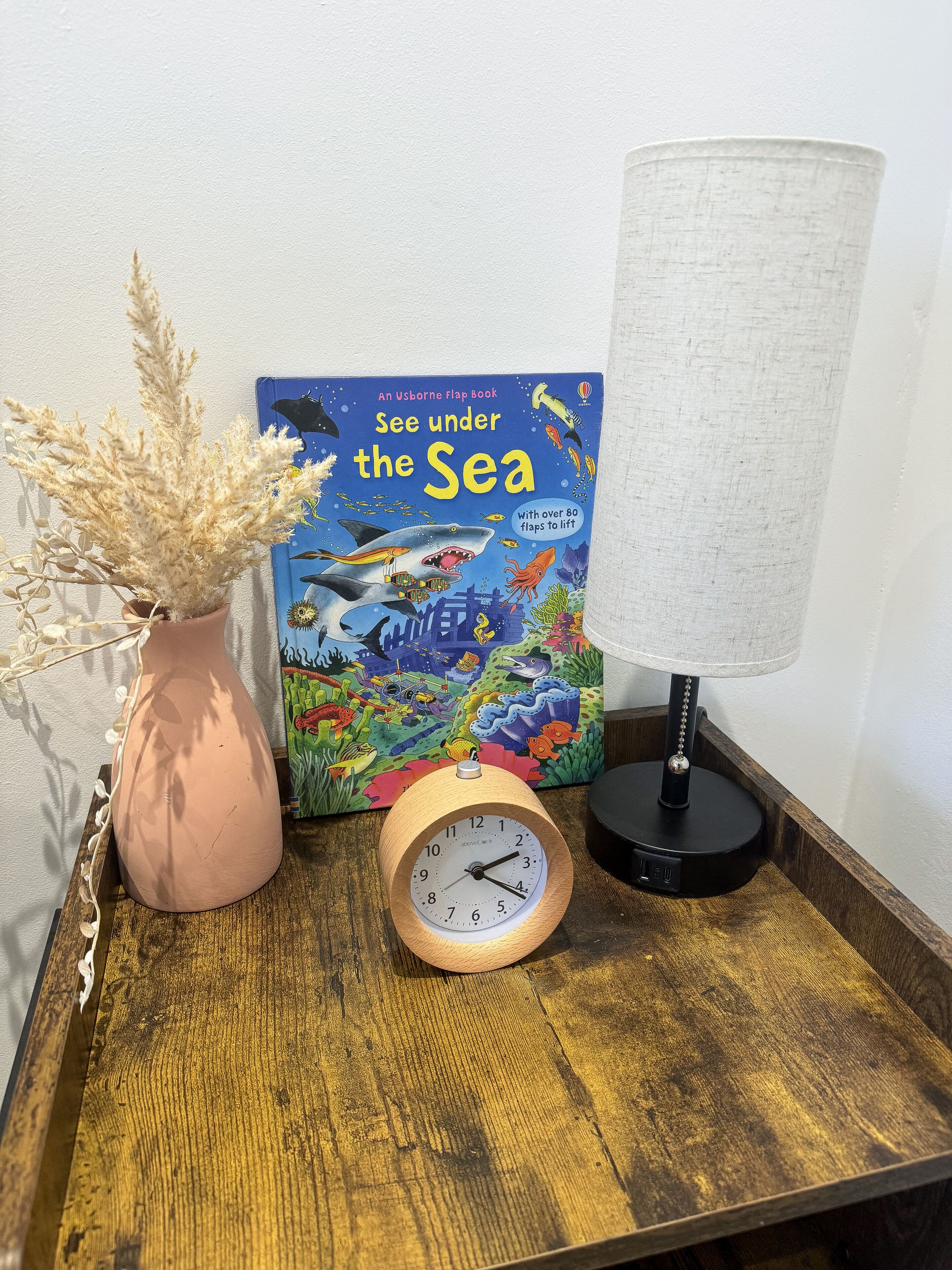 A wooden bedside table with a clock, a book titled "See under the Sea," and a vase with dried flowers.