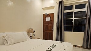 Una cama bien hecha con una colcha blanca, una puerta de madera, una ventana con cortinas y una lámpara de noche.