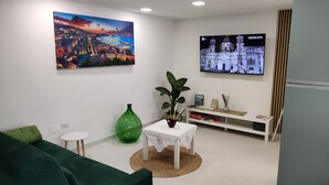 A living room with a green sofa, a small white table with a plant, a TV mounted on the wall, and a painting of a cityscape. - Originale, Elegante e Confortevole Location nei Pressi Della Città Antica (Pompei)