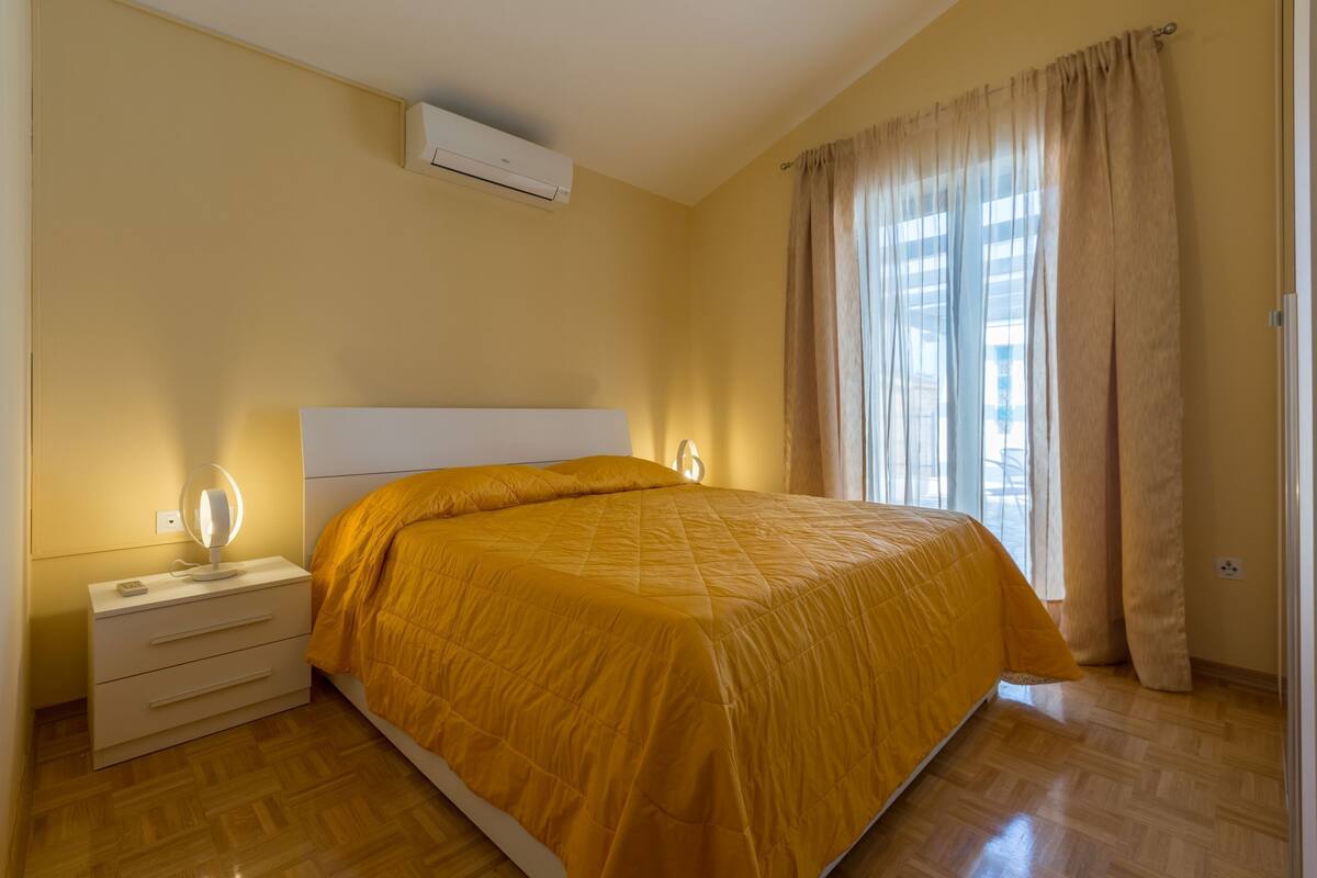 Quarto com cama, mesa de cabeceira, abajur e janela com cortinas.