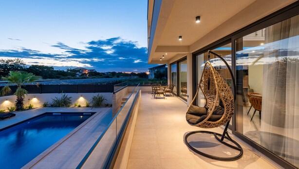 Een modern terras buiten met een zwembad, een hangstoel en een overdekte eethoek van glas.