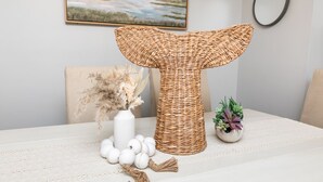 Una mesa con un centro de mimbre con forma de cola de ballena, un florero con flores secas y una pequeña planta en maceta.