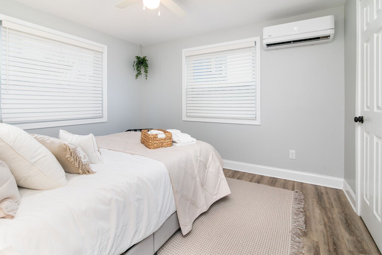 A bedroom with two beds, white bedding, a woven basket, and an air conditioning unit on the wall.