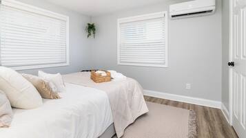 A bedroom with two beds, white bedding, a woven basket, and an air conditioning unit on the wall.