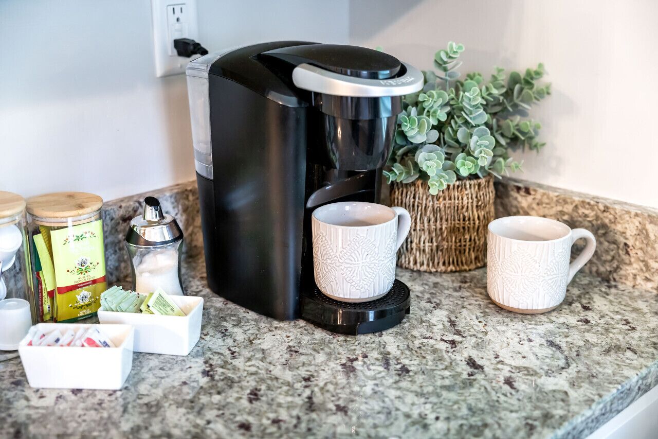 A coffee maker on a granite countertop with two white cups, a small jar, and a box of tea bags.