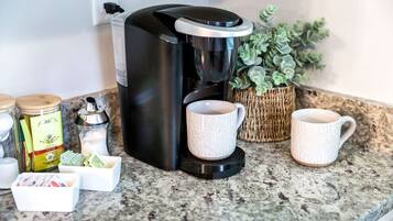 A coffee maker on a granite countertop with two white cups, a small jar, and a box of tea bags.