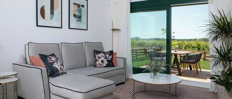 Un salon moderne avec un canapé gris, une table basse ronde et une vue sur un jardin luxuriant, visible à travers une porte-fenêtre coulissante.