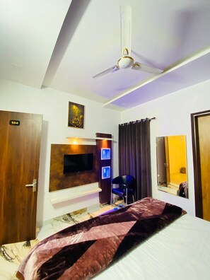A hotel room with a bed, a ceiling fan, a TV mounted on the wall, a mirror, and a door with a number sign. - Hotel Azaara (Patiala)