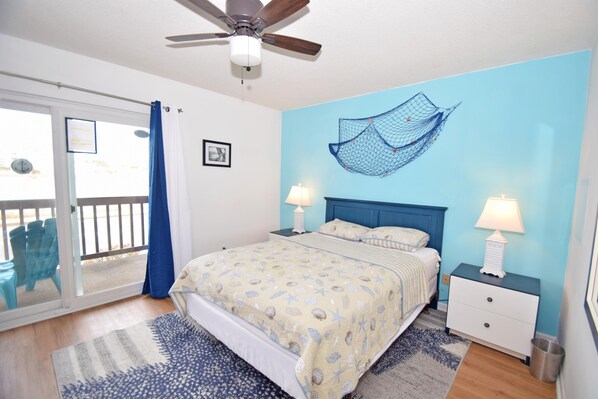 Quarto com cama, duas mesas de cabeceira com luminárias, ventilador de teto e vista através de uma porta deslizante para área externa com cadeiras.