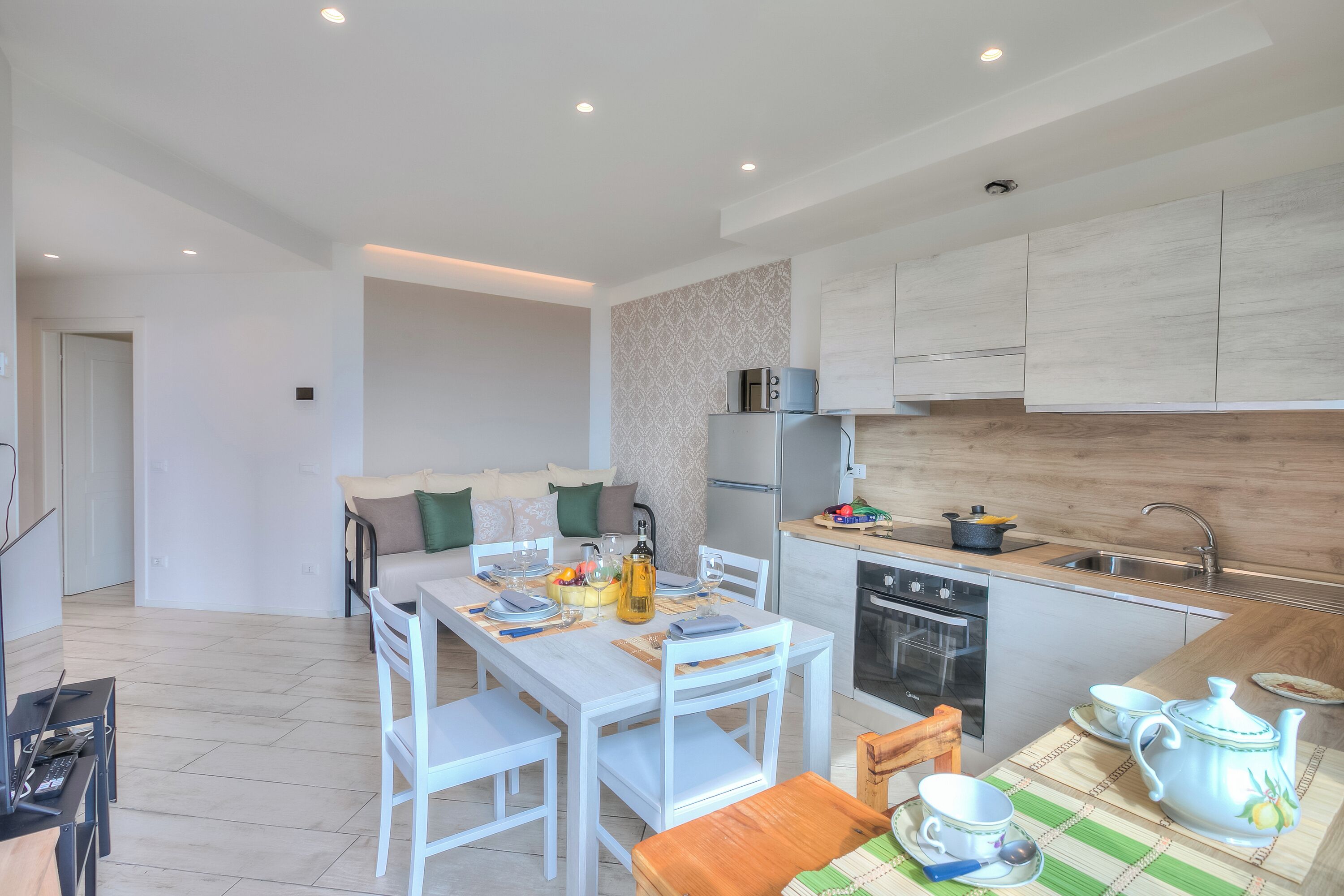 Una cocina moderna con armarios blancos, una zona de comedor con mesa y sillas blancas, y un pequeño rincón de estar con sofá y cojines.